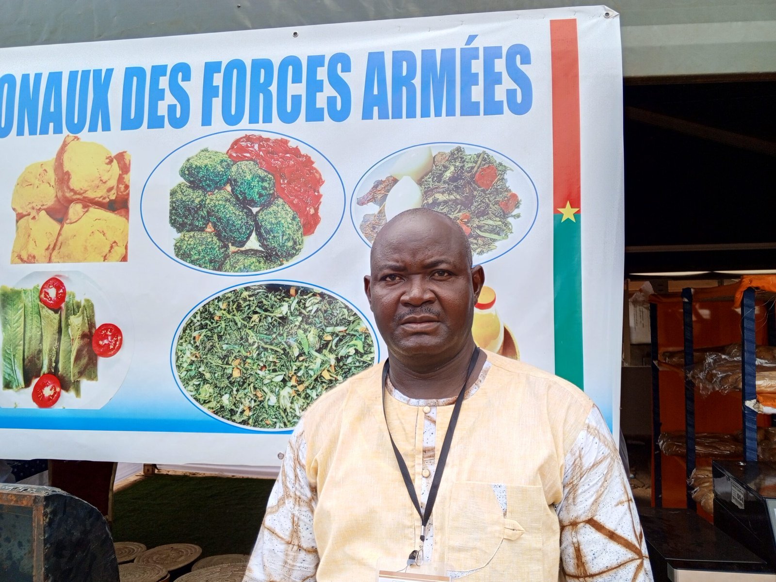 L'adjudant-chef Oumarou OUEDRAOGO au stand des Forces armées devant le panneau des spécialités culinaires.